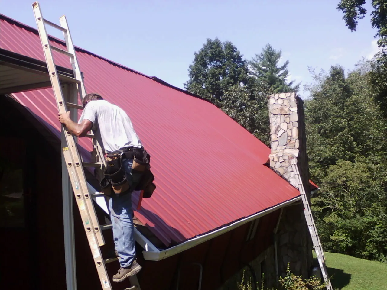Roofer on a ladder beside a red metal roof and stone chimney — Soffit & Fascia Repair by Roofing Brothers in Iola